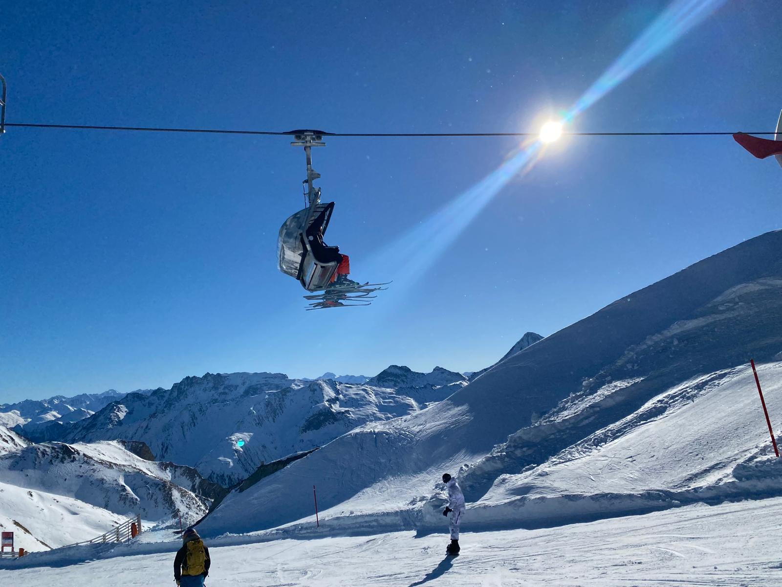 Chairlift crossing a deep blue sky above a snowy mountain panorama, with two skiers on the piste below
